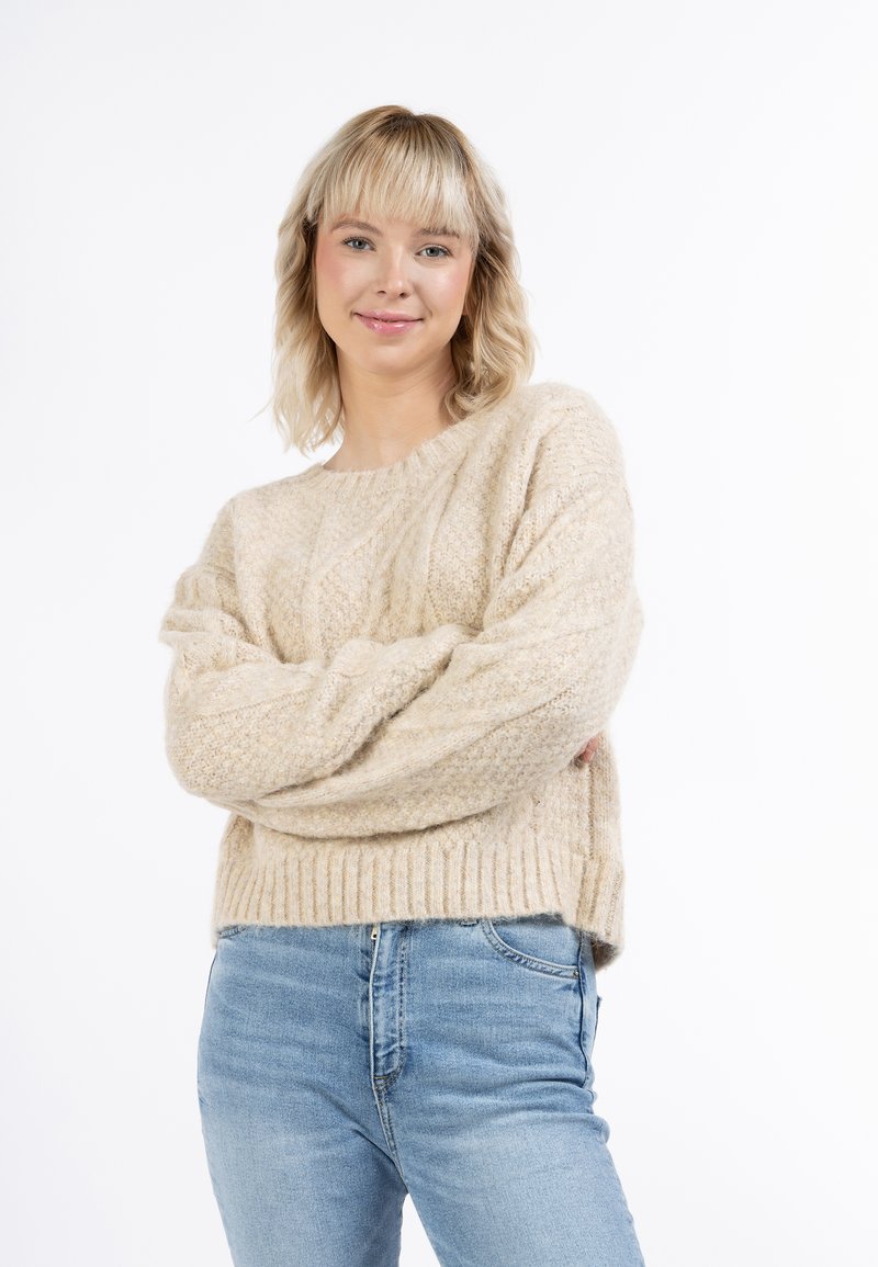 Beige kötött pulóver bordás szegéllyel és textúrált mintával, világoskék farmernadrággal párosítva. Az anyag lágy és meleg hatású.