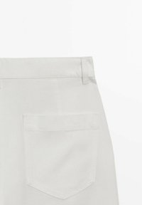 Vue arrière d'un pantalon beige clair montrant une seule poche arrière et des passants pour ceinture sur un fond blanc uni.