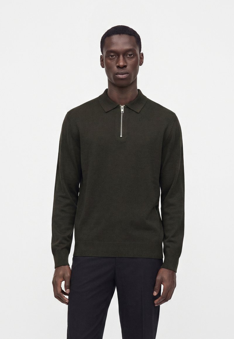 Homme portant un polo à manches longues vert foncé avec fermeture éclair et pantalon foncé, debout devant un fond blanc uni.