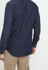 Hombre vestido con una camisa de mezclilla de manga larga azul oscuro y pantalones cargo beige, visto desde atrás contra un fondo sencillo.