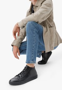 Čierne vysoké tenisky s hladkým koženým povrchom a gumovou podrážkou, doplnené svetlomodrými džínami a béžovou semišovou bundou.