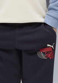 Ciemnoniebieskie dresy wykonane z miękkiego materiału mają czerwono-białe logo Pumy oraz graficzny wzór na lewej kieszeni udowej.