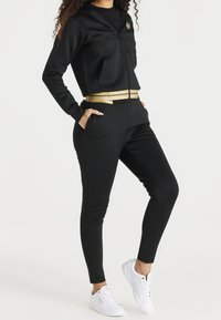 Zwarte hoodie met ritssluiting en gouden accenten, voorzien van zakken, gecombineerd met zwarte taps toelopende broek met een gouden tailleband. Witte sneakers maken de outfit compleet.