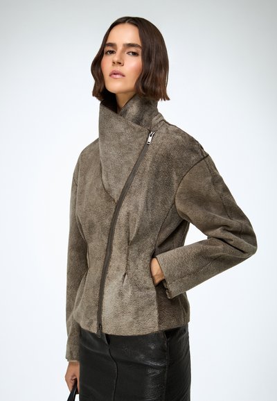 Barna szarvasbőr dzseki magas gallérral és aszimmetrikus cipzárral. Laza fazon és puha textúra jellemzi, fekete bőrszoknyával párosítva.