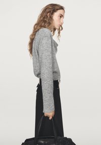 Vrouw met golvend haar draagt een grijze trui, zwarte brede broekspijpen, een brede zwarte riem en houdt een grote zwarte handtas vast tegen een effen achtergrond.