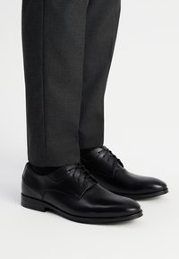 Czarne skórzane buty formalne o gładkim wykończeniu, z wiązaniem i okrągłym noskiem. Połączone z ciemnoszarymi, dopasowanymi spodniami.