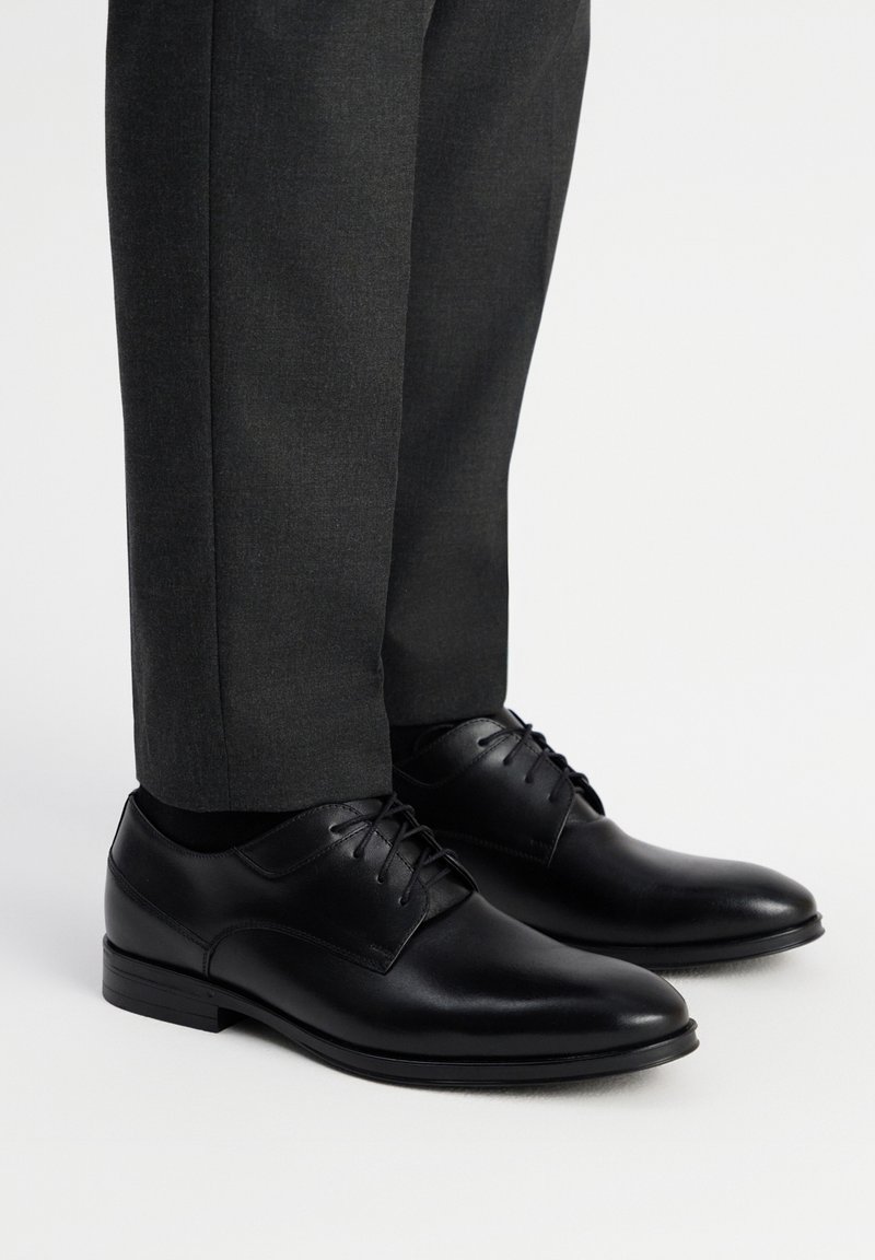 Czarne skórzane buty formalne o gładkim wykończeniu, z wiązaniem i okrągłym noskiem. Połączone z ciemnoszarymi, dopasowanymi spodniami.