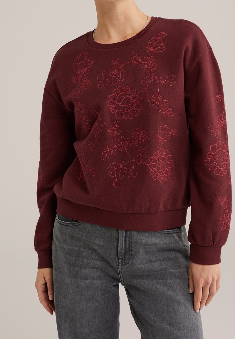 Sweatshirt bordeaux avec broderie florale rouge porté avec un jean gris, montrant le haut du corps et une coupe décontractée.