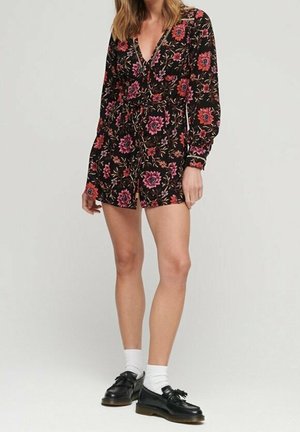 Zwarte bloemenromper met lange mouwen, versierd met roze en rode bloemen, een diepe V-hals en een getailleerde taille. Gecombineerd met zwarte schoenen en witte sokken.