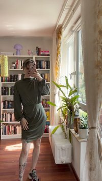 Olivově zelené šaty s dlouhým rukávem a stahovaným pasem, vyrobené z měkkého materiálu. Modelka má na sobě vzorované boty a stojí u knihoven a rostlin.