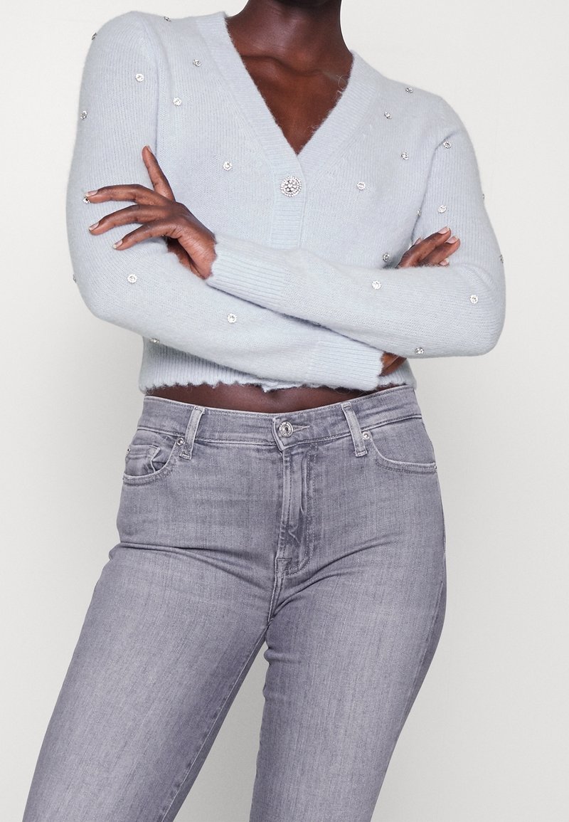 Personne portant un cardigan court bleu clair orné d'accents en strass et un jean gris taille haute, les bras croisés sur la poitrine.