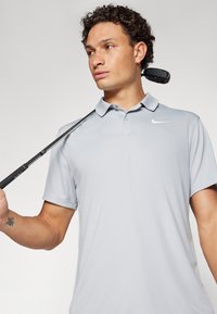 Svijetlosiva polo majica s ovratnikom i kratkim rukavima, s logotipom Nike. Model drži golf palicu preko ramena.