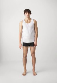 Jauns vīrietis stāv basām kājām uz vienkārša fona, ģērbies baltā bez piedurknēm krekla un melnos šortos, vērsts pret priekšu ar atslābinātām rokām.