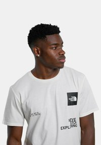 Camiseta blanca de algodón con mangas cortas, que presenta un parche de logo negro y texto impreso en negro en la parte frontal. Cuello redondo, corte relajado.