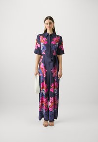 Mørkeblå blomster maxi-kjole med korte ærmer, prydet med rosa og lilla blomster. Har et bånd i midjen og er kombinert med en hvit håndveske.