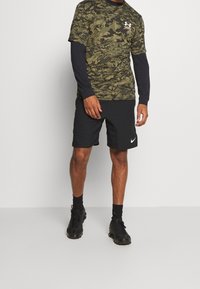 Camisa de manga corta de camuflaje verde combinada con pantalones cortos deportivos negros, junto con zapatillas deportivas negras y calcetines de longitud hasta el tobillo.