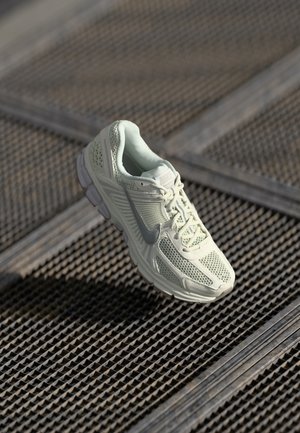 Hvid Nike løbesko med netdesign, hængende over tekstureret metal gittergulv.