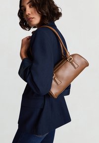 Borsa a tracolla in pelle marrone con una forma strutturata, tracolla regolabile e dettagli in metallo color oro, indossata sulla spalla sopra un blazer blu scuro.