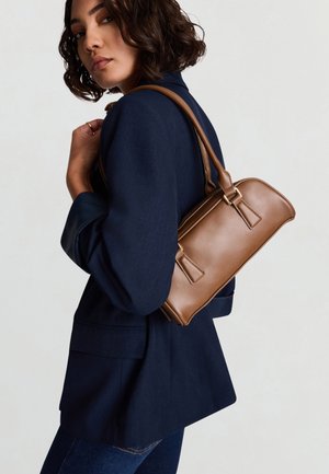 Braune Lederhandtasche mit strukturierter Form, verstellbarem Riemen und goldfarbenen Beschlägen, getragen über der Schulter zu einem Marineblazer.