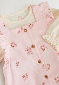 Vestido de peto de pana rosa con estampado floral, que cuenta con botones de madera y detalles de volantes en los hombros. Incluye una blusa de manga larga color crema.