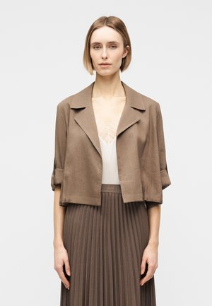 Γυναίκα με κοντά ξανθά μαλλιά που φοράει ένα ανοιχτό καφέ κοντό μπουφάν πάνω από λευκό δαντελένιο τοπ και πλισέ καφέ φούστα.