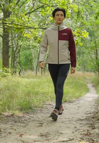 Lekka, beżowa i bordowa kurtka trekkingowa z wysokim kołnierzem, w komplecie z czarnymi legginsami i solidnymi brązowymi butami trekkingowymi, idąca po ścieżce gruntowej.
