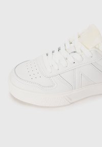 White sneaker with a textured rubber sole, featuring round perforations, flat laces, and side design accents in a minimalist style.