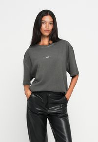 T-shirt cinza oversized com gola redonda e mangas curtas, apresentando um pequeno logótipo branco. Combinado com calças pretas brilhantes.