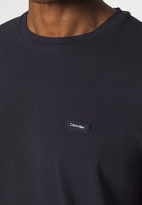 Gros plan sur une personne portant un t-shirt à col rond bleu marine foncé avec un petit patch rectangulaire du logo Calvin Klein sur la poitrine.