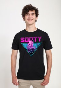 Černé bavlněné tričko s grafickým designem, který zobrazuje fialového medvídka a slovo "SOOTY" tučným růžovým písmem uvnitř tyrkysového trojúhelníku.