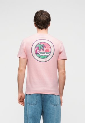 Muž nosiaci ružové tričko s kruhovou grafikou palmových stromov, vĺn a západu slnka, v páre s voľnými modrými džínsami, otočený chrbtom.