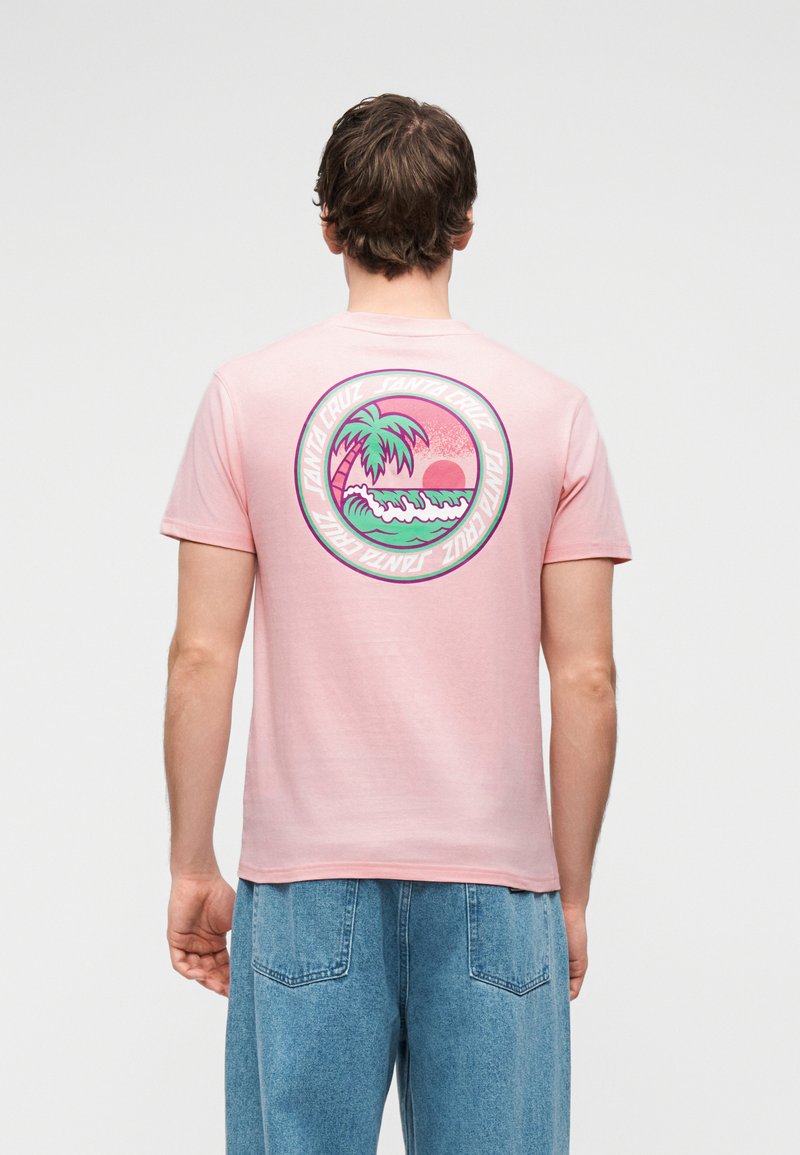 Άνδρας που φοράει ροζ μπλουζάκι με κυκλικό σχέδιο φοίνικες, κύματα και ηλιοβασίλεμα, συνδυασμένο με φαρδιά μπλε τζιν, με την πλάτη γυρισμένη.