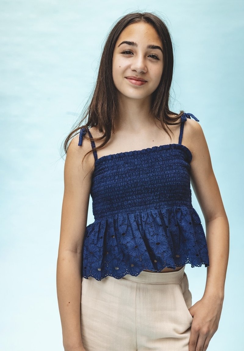 Marineblauwe smocked crop top met dunne bandjes en een gegolfde zoom, versierd met geborduurde bloemenpatronen. Gecombineerd met lichtgekleurde pantalons.