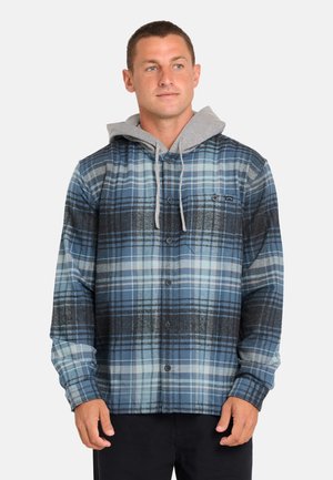 Mann trägt ein blau-grau kariertes Hemd über einem grauen Kapuzenpullover und steht vor einem einfarbigen hellen Hintergrund.