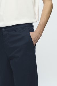Dark navy chinos with a smooth texture, featuring a button closure, side pockets, and a tailored fit, paired with a light-colored top.