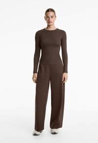 Donna in piedi di fronte, che indossa un top marrone a maniche lunghe aderente, pantaloni marroni a gamba larga e sneakers bianche su uno sfondo bianco.