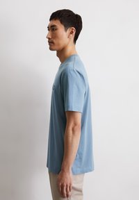 Blauwe katoenen T-shirt met korte mouwen, ronde hals en rechte snit, gedragen door een persoon die naar links kijkt, tegen een effen achtergrond.