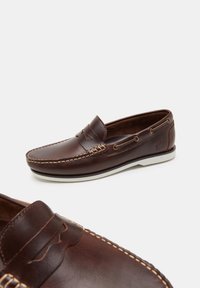Brown leather loafers with white rubber soles, decorative stitching, and side lacing detail. Smooth texture with subtle branding on heel.