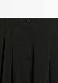 Gros plan sur une jupe plissée en tissu noir avec un bouton et une fermeture éclair dissimulée à la taille.