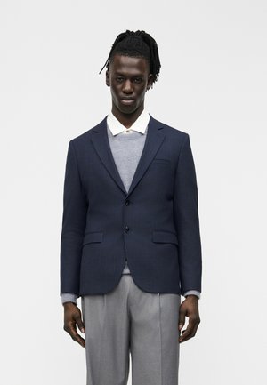 Homme aux cheveux tressés portant un blazer bleu marine sur un pull gris et une chemise blanche à col, assorti à un pantalon gris, debout devant un fond blanc.