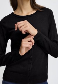 Cardigan negru tricotat cu guler rotund, nasturi în față și mâneci lungi. Prezintă o textură netedă și o siluetă ajustată.