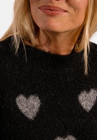 Maglione nero a maglia con motivi a cuore grigi. Collo rotondo e texture morbida, che mostra un primo piano dei cuori sul davanti.