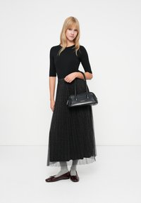 Kvinne iført svart topp, lang mønstret svart skjørt, grå sokker, burgunder leiligheter, holder en svart veske mot en hvit bakgrunn.