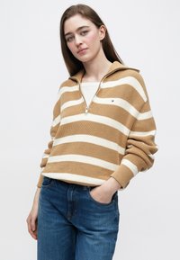 Béžovo-biely pruhovaný pulóverový sveter so zipsovým golierom a rebrovanou štruktúrou, v kombinácii s modrými rifľami.