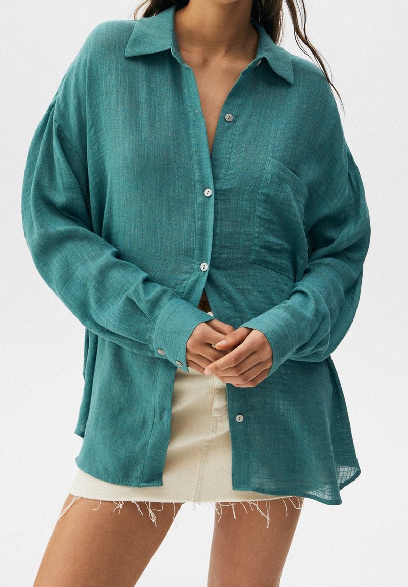 Woman wearing a teal oversized button-up shirt with a chest pocket and a short beige skirt with frayed hem, hands clasped in front.