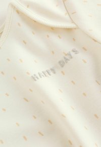 Világos krémszínű, pamut body kis, pontozott, mézszínű mintákkal. "HAPPY DAYS" felirattal halványszürke betűkkel. Rövid ujjú és bordázott nyakkivágású.