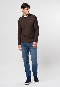 Muž nosiaci tmavohnedú polokošeľu s dlhým rukávom, modré džínsy a čierne tenisky stojaci pred jednofarebným svetlým pozadím.