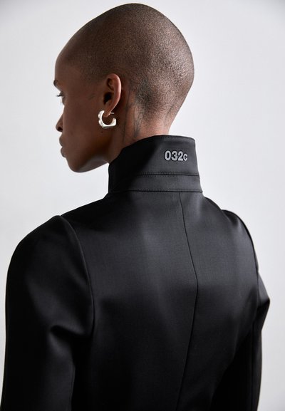 Black structured jacket with high collar and a visible brand tag "032c" on the neck. Features a smooth texture and tailored fit.