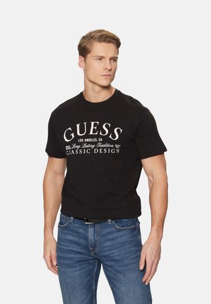 Camiseta de algodón negra con texto y elementos de diseño del logo "GUESS" en blanco. Con mangas cortas, escote redondo, combinada con unos vaqueros de denim azul.
