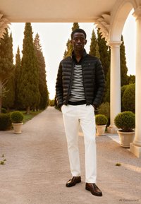 Jeune homme debout sur un chemin de gravier bordé de hauts cyprès, portant une doudoune noire, un pull rayé, un pantalon blanc et des chaussures en cuir marron.